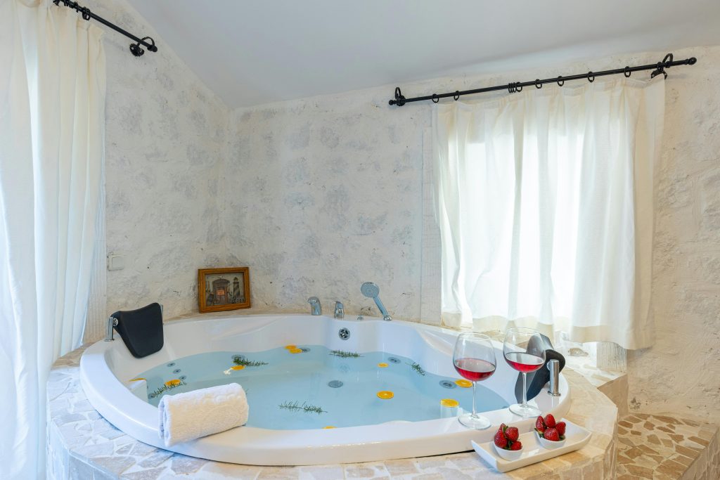 Corner jacuzzi tub with flower petals, wine glasses, and towels in a quiet stone bathroom with natural light